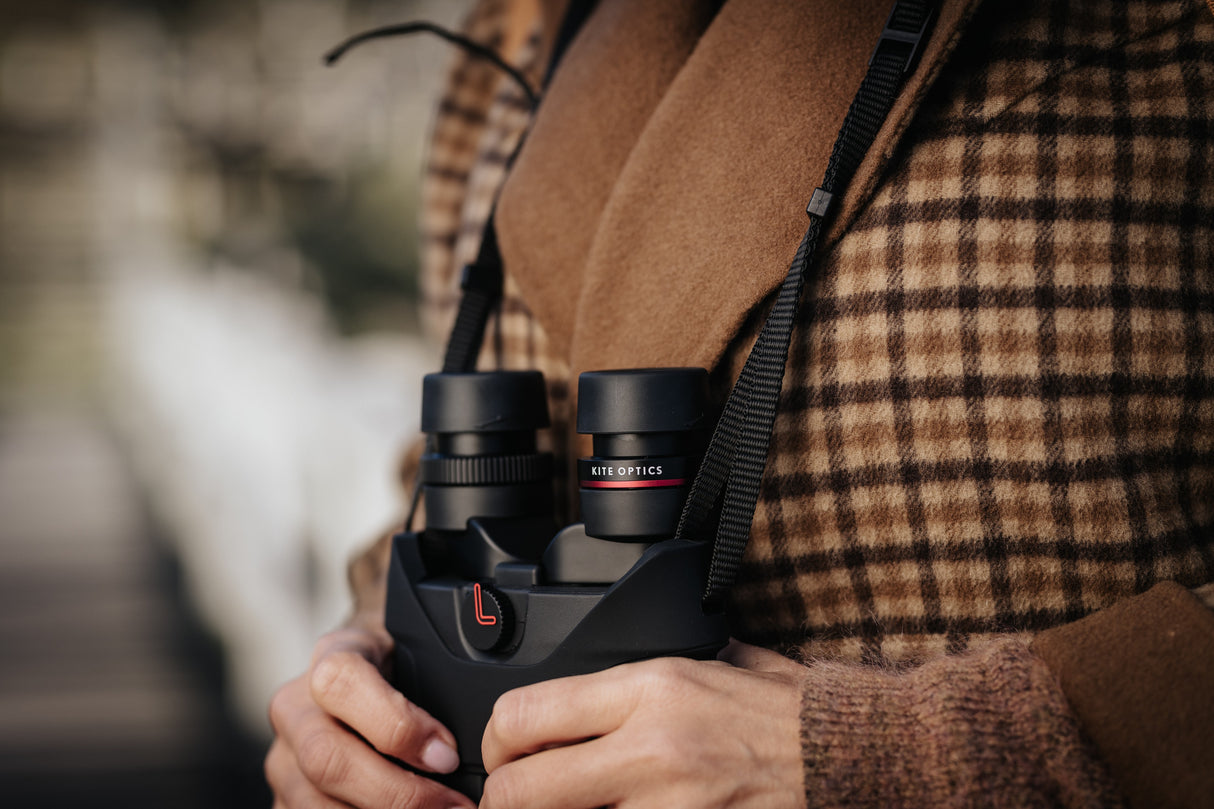 Kite Optics APC 42 stabilized binoculars hanging from neoprene neckstrap, close-up showing eyepieces and on/off switch