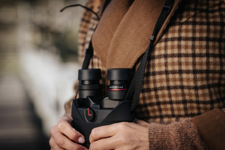 Kite Optics APC 42 stabilized binoculars hanging from neoprene neckstrap, close-up showing eyepieces and on/off switch